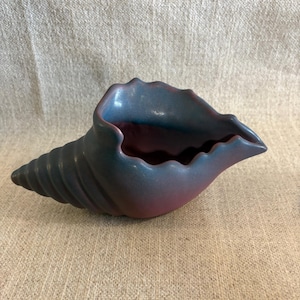 May include: A ceramic seashell-shaped decorative bowl. The bowl features a gradient of colors, transitioning from a deep blue to a soft pink. The spiral end is a darker blue, while the interior has a pink hue. The bowl is placed on a neutral-toned fabric.