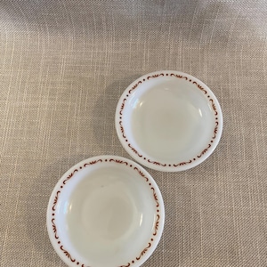 May include: Two small, white, round bowls with a brown floral design around the rim. The bowls are empty and sit on a neutral-coloured, textured surface. The bowls are likely for serving dips or small portions.