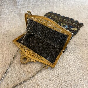 May include: An antique black mesh purse with a gold-toned filigree frame and a delicate chain strap. The purse is open, revealing the intricate mesh interior. The top flap is decorated with colorful beads.