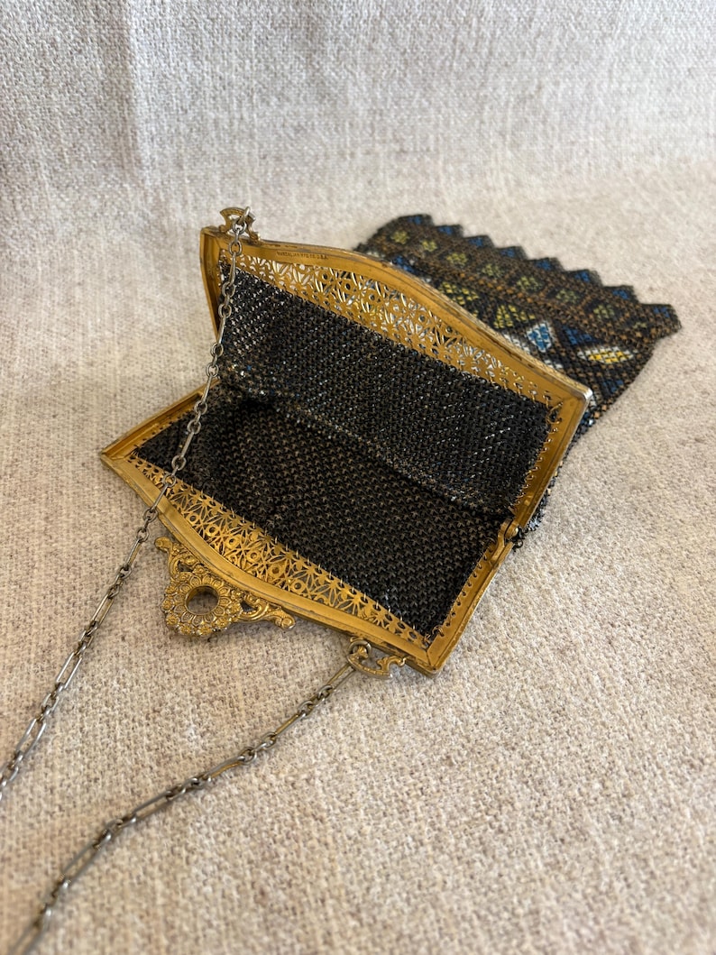 May include: An antique black mesh purse with a gold-toned filigree frame and a delicate chain strap. The purse is open, revealing the intricate mesh interior. The top flap is decorated with colorful beads.