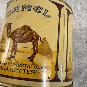 May include: Vintage Camel cigarettes tin with a tan and brown color scheme. The tin features a camel, pyramids, and palm trees. Text on the tin reads "Camel" and "Domestic Blend Cigarettes".