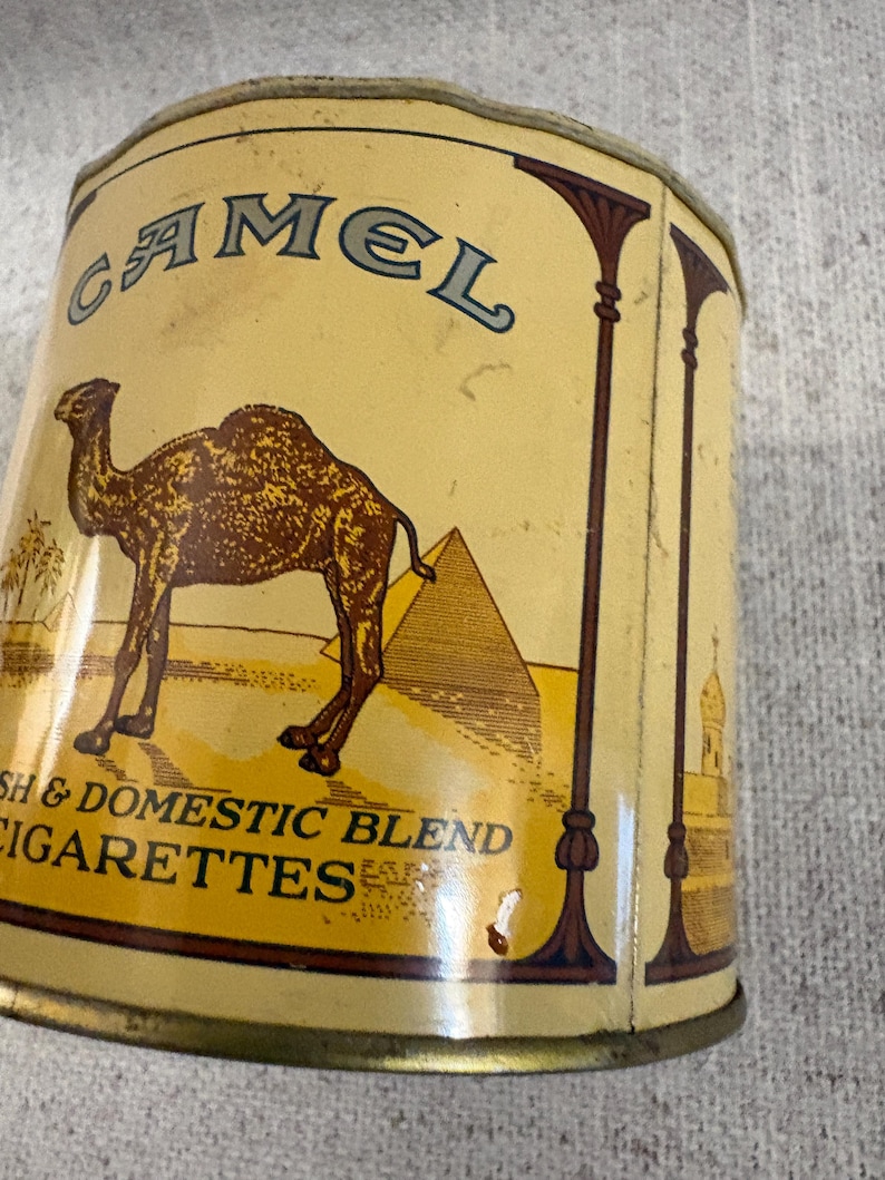 May include: Vintage Camel cigarettes tin with a tan and brown color scheme. The tin features a camel, pyramids, and palm trees. Text on the tin reads "Camel" and "Domestic Blend Cigarettes".