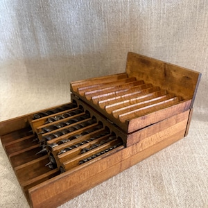 May include: A tiered wooden storage rack featuring multiple horizontal grooves designed to hold metal auger drill bits. The rack is constructed from light brown wood with a visible grain, organized into two levels to accommodate various tool sizes.