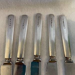 May include: Set of five silver butter knives with Art Deco style handles. Each knife is engraved with a different herb name: sage, mint, chives, basil, and fennel. The blades are rectangular and the handles are rectangular with decorative geometric patterns.