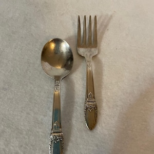 Puede incluir: Un juego de cuchara y tenedor de plata. La cuchara tiene un cuenco redondeado y un mango decorativo. El tenedor tiene cuatro púas y un mango a juego. Ambos utensilios tienen detalles florales ornamentados en la base del mango.