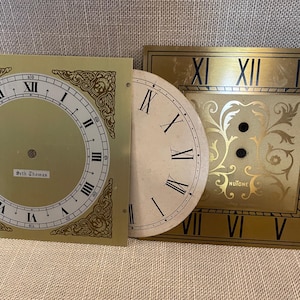 May include: Three vintage clock faces in gold and cream. One is square with Roman numerals and the name "Beth Thomas." Another is a half-circle with black Roman numerals. The third is rectangular with Roman numerals and the word "NuTone."