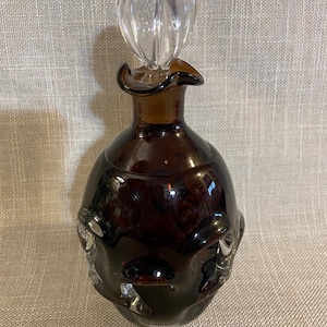 Puede incluir: Un frasco de perfume de vidrio ámbar oscuro con un tapón de vidrio transparente y acanalado. La botella tiene forma bulbosa con elementos decorativos texturizados cerca de la base. El tapón es de forma ovalada y tiene un diseño acanalado.