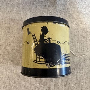 Puede incluir: Una lata de metal vintage con tapa y base negras. La lata presenta una silueta de una persona tejiendo en una mecedora, con una escalera y elementos decorativos. El fondo es amarillo pálido.
