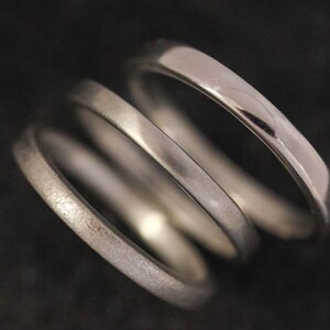 May include: Three silver rings are stacked together, each with a different texture and finish. The top ring is highly polished, reflecting light, while the middle ring has a matte finish. The bottom ring has a textured, almost hammered appearance. The rings are set against a dark background.