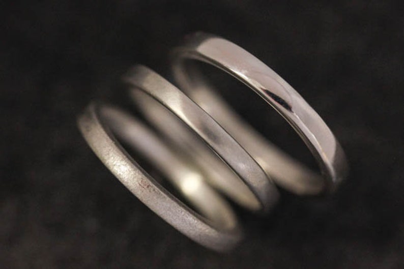 May include: Three silver rings are stacked together, each with a different texture and finish. The top ring is highly polished, reflecting light, while the middle ring has a matte finish. The bottom ring has a textured, almost hammered appearance. The rings are set against a dark background.