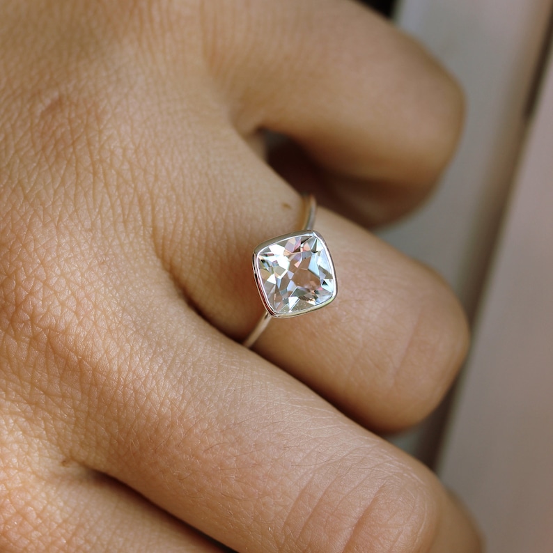 May include: A silver ring featuring a square, clear gemstone. The gemstone is set in a bezel setting, and the ring is on a finger. The ring's band is thin and made of silver.