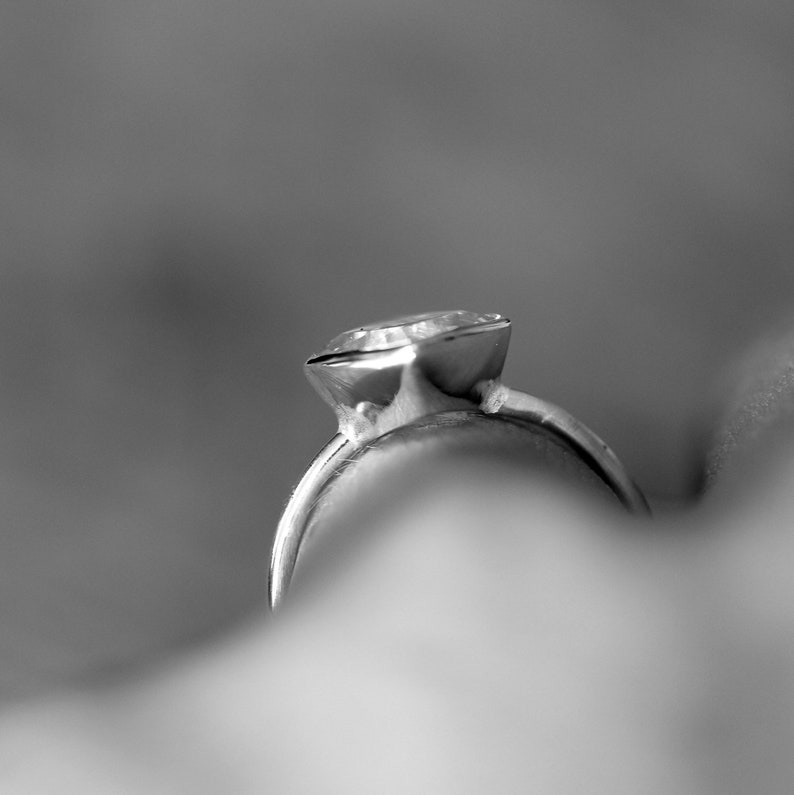 May include: Close-up of a silver ring with a large, clear gemstone. The ring's band is smooth and curved, leading up to a bezel setting that holds the sparkling stone. The background is blurred, emphasizing the ring's details.