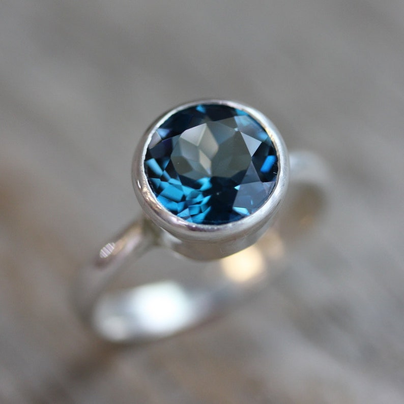 May include: A close-up of a silver ring featuring a large, round, faceted, deep teal gemstone. The ring's band is smooth and polished, and the gemstone is set in a bezel setting. The ring is displayed against a blurred wooden background.