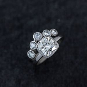 May include: A silver engagement ring set with a large, square-cut diamond and a matching band. The engagement ring features a halo of smaller round diamonds. The rings are set against a dark background.