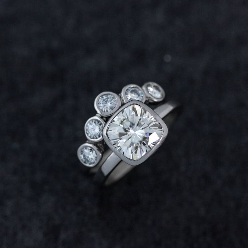 May include: A silver engagement ring set with a large, square-cut diamond and a matching band. The engagement ring features a halo of smaller round diamonds. The rings are set against a dark background.