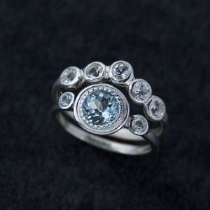 May include: Two silver rings with gemstones. The top ring has six small, clear gemstones. The bottom ring features a large, round, light blue gemstone surrounded by smaller clear gemstones. The rings are set against a dark background.