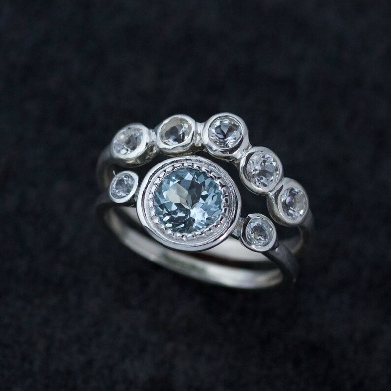 May include: Two silver rings with gemstones. The top ring has six small, clear gemstones. The bottom ring features a large, round, light blue gemstone surrounded by smaller clear gemstones. The rings are set against a dark background.