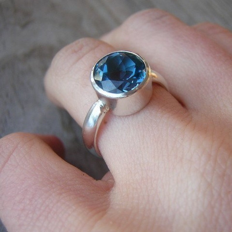May include: A silver ring with a large, round, faceted, deep blue gemstone. The ring is set in a simple bezel setting, and the band is smooth and polished. The ring is on a person's finger.