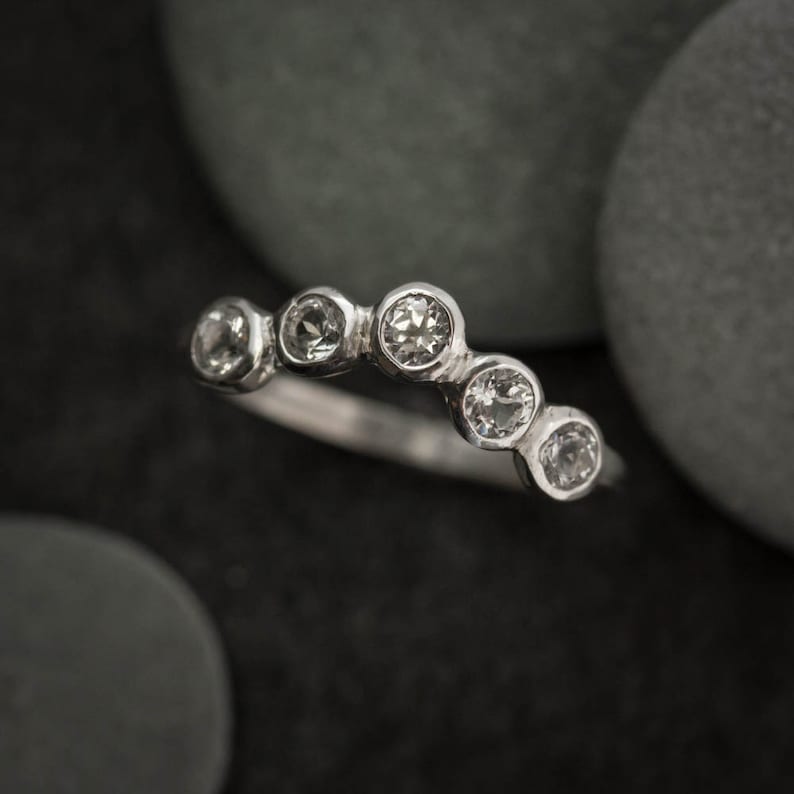 May include: A silver ring with five round, clear gemstones set in a curved design. The ring is displayed against a dark background with smooth, gray stones.