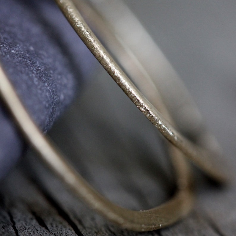 Op de afbeelding: Close-up van twee gouden hoepels, een iets groter dan de andere, met een getextureerd oppervlak. De hoepels liggen op een grijs, getextureerd oppervlak.