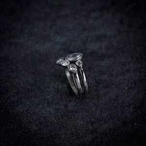 May include: A silver ring with a cluster of gemstones. The ring features a large, oval-shaped, light blue gemstone at the top, surrounded by smaller, clear gemstones. The band is split into two parallel lines.