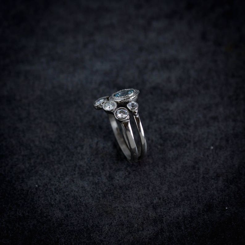 May include: A silver ring with a cluster of gemstones. The ring features a large, oval-shaped, light blue gemstone at the top, surrounded by smaller, clear gemstones. The band is split into two parallel lines.