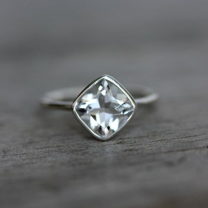 May include: A silver ring with a square, clear gemstone. The gemstone is set in a bezel setting, and the band is thin and polished. The ring is displayed on a wooden surface.