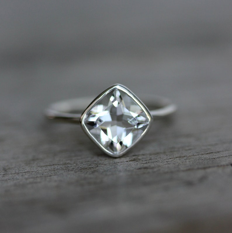 May include: A silver ring with a square, clear gemstone. The gemstone is set in a bezel setting, and the band is thin and polished. The ring is displayed on a wooden surface.