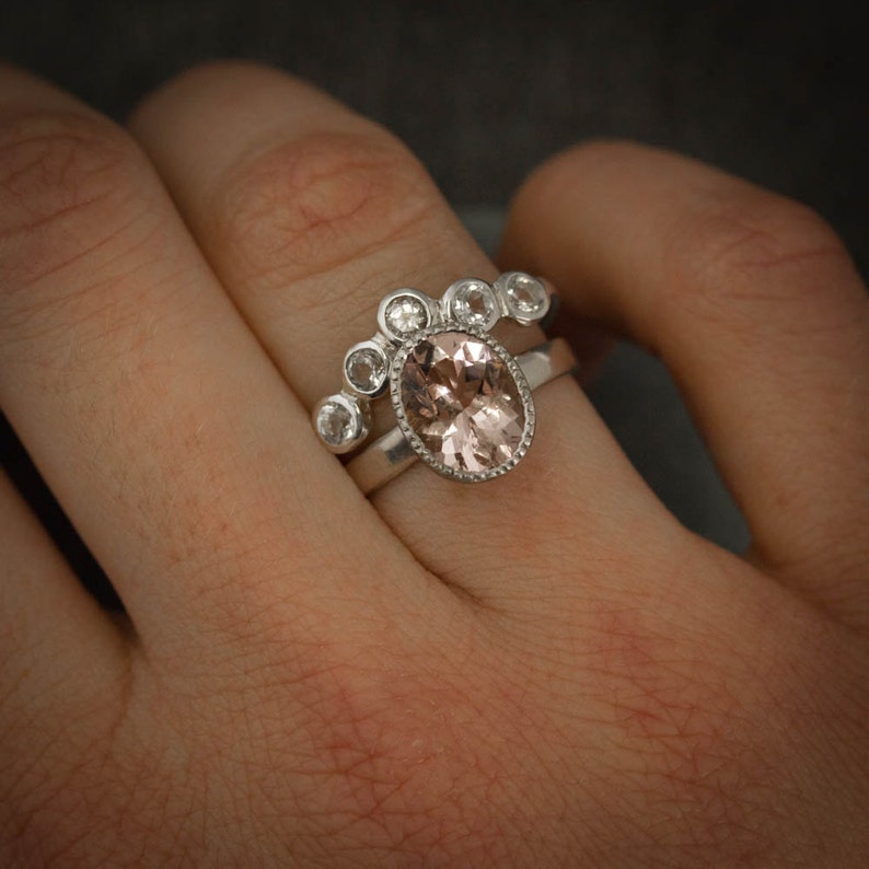 May include: A silver ring set featuring a large oval peach-colored gemstone with a textured bezel. The ring is paired with a curved band adorned with smaller, clear gemstones. The rings are displayed on a finger.