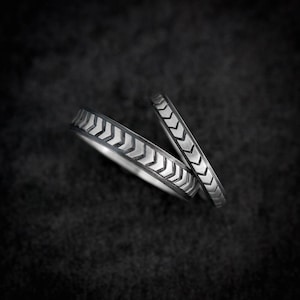 May include: Two silver rings with a chevron pattern. The rings are set against a dark background, highlighting the intricate design. The chevron pattern is repeated around the bands, creating a modern and geometric look.