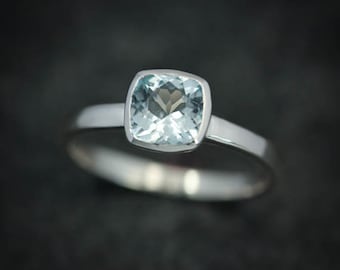 May include: A silver ring featuring a square, light blue gemstone. The gemstone is set in a bezel setting, and the band is a simple, polished silver. The ring is displayed against a dark background.