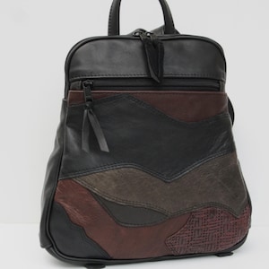 May include: A multi-textured backpack featuring a patchwork design of black, dark brown, and burgundy leather panels. The bag includes a top handle, adjustable straps, and a front zippered pocket with black zipper pulls and matching leather tabs.