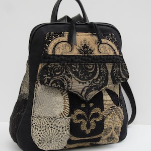 May include: A black backpack featuring a front pocket flap with a textured, woven pattern in black and metallic gold. The bag includes a top handle, adjustable shoulder straps, and a zippered main compartment with black leather-like trim.