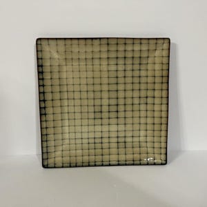 May include: Square decorative plate with a grid pattern. The plate has a beige background with a dark grid overlay. The edges of the plate are a darker brown color. The plate is sitting on a white surface.