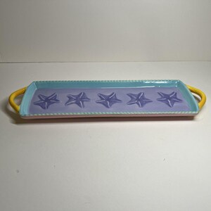May include: A rectangular ceramic serving tray with a pastel color scheme. The tray features a light pink base, a lavender interior with five starfish, a turquoise rim, and yellow handles. The rim has a rope-like design.