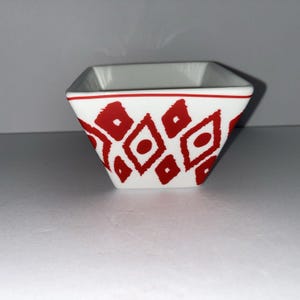 Puede incluir: Un cuenco cuadrado blanco con un patrón geométrico rojo. El cuenco tiene un borde rojo y una serie de formas de diamante rojas con un punto en el centro. El cuenco está vacío y descansa sobre una superficie blanca.