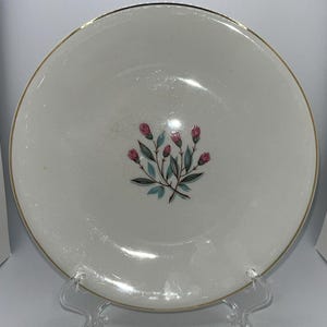 Puede incluir: Un plato de cerámica blanca con borde dorado y diseño floral. El diseño presenta capullos de flores rosas, hojas turquesas y tallos verdes. El plato se exhibe sobre un soporte de plástico transparente.