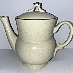 May include: Cream-colored ceramic teapot with a speckled pattern. Features a curved spout, a rounded body, and a handle. The lid has a decorative handle. A classic piece for serving tea.
