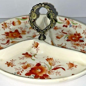 Pode incluir: Travessa ovalada de três secções com um padrão floral em laranja, vermelho e verde sobre um fundo creme. Possui uma pega decorativa em metal. A travessa é provavelmente feita de cerâmica ou porcelana.