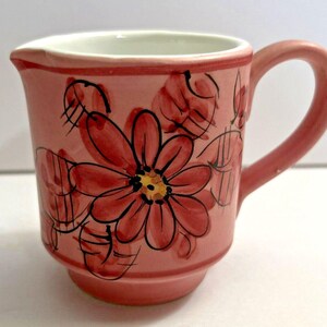 May include: A pink ceramic creamer with a handle and a spout. The creamer is decorated with large red flowers with black outlines and yellow centers. The rim and handle are also pink.