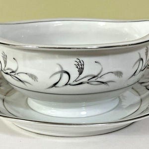 May include: White gravy boat with a matching saucer. The set features a silver trim and a black and silver wheat stalk design. The gravy boat has a curved handle and a spout for easy pouring. A classic addition to any dining table.