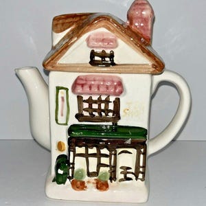 May include: A white ceramic teapot shaped like a house, with a brown roof and a pink chimney. The teapot has a handle and a spout. The front of the teapot has a painted design of a shop with the text "Tea Shop".