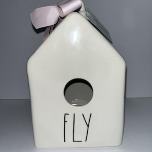 Puede incluir: Una pajarera de cerámica blanca con una abertura redonda y la palabra "FLY" en letras negras. Una cinta rosa claro está atada en la parte superior. La pajarera es un artículo decorativo.