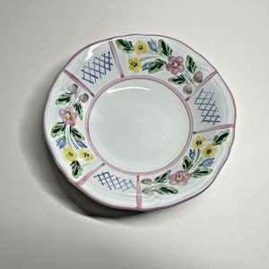 May include: White ceramic plate with a scalloped edge, decorated with a floral pattern in yellow, pink, and blue. The plate has a pink border and blue crosshatch designs. A decorative piece for serving food.