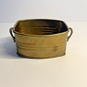 Op de afbeelding: Een vintage messing plantenbak met een rechthoekige vorm, afgeronde hoeken en kleine handgrepen. Het messing heeft een warme, verouderde patina. Het ontwerp van de plantenbak suggereert dat het een antiek of vintage stuk is. Afmetingen: ongeveer 15 cm lang, 10 cm breed en 8 cm hoog.