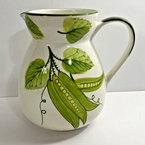 Op de afbeelding: Crème keramische kan met een groene rand en handvat. De kan is versierd met groene peulen en bladeren. Het handvat en de rand zijn groen geschilderd. De kan heeft een klassieke, afgeronde vorm.