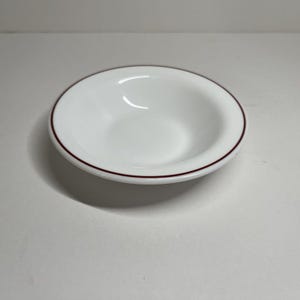 Könnte beinhalten: Eine kleine, weiße Keramikschale mit runder Form und einem dünnen, dunkelroten Rand. Die Schale ist leer und steht auf einer weißen Oberfläche. Die Schale ist wahrscheinlich zum Servieren von Dips, Saucen oder kleinen Portionen gedacht.