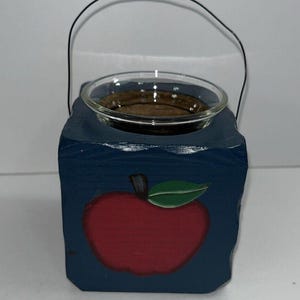 May include: A blue wooden candle holder with a red apple design and a clear glass dish. The candle holder has a black wire handle. The apple has a green leaf and a black stem.