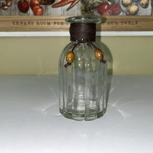 May include: A clear glass vase with a brown rope detail around the neck and two wooden beads. The vase has a ribbed design and a flared top. The background features a wooden sign with the text "THERE'S ROOM FOR YOUR TABLE".