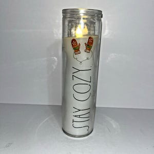 以下が含まれることがあります： 点灯したLED炎のある透明なガラスのキャンドル。キャンドルには、黒で「STAY COZY」という言葉とミトンのペアが書かれた白いラベルが付いています。ミトンは茶色で、赤と緑のアクセントが付いています。